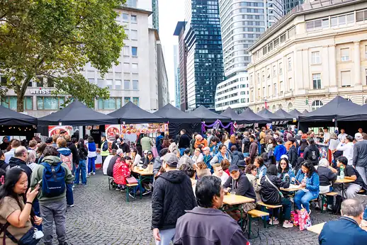Voller Food-Court mit langen Tischreihen und Zelten, dahinter die Skyline