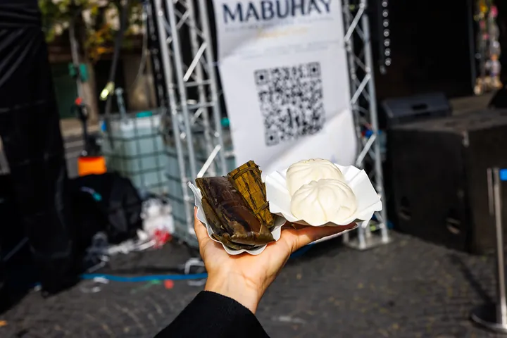 Eine Hand hält Suman und Siopao vor das MABUHAY-Banner an der Bühne