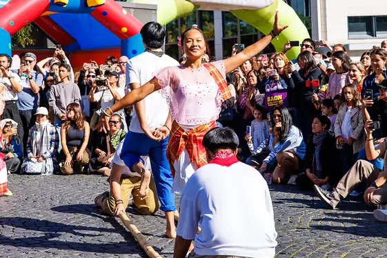 Tinikling: Tänzerin in rosa Tracht breitet die Arme aus, vorne knien die Bambusträger